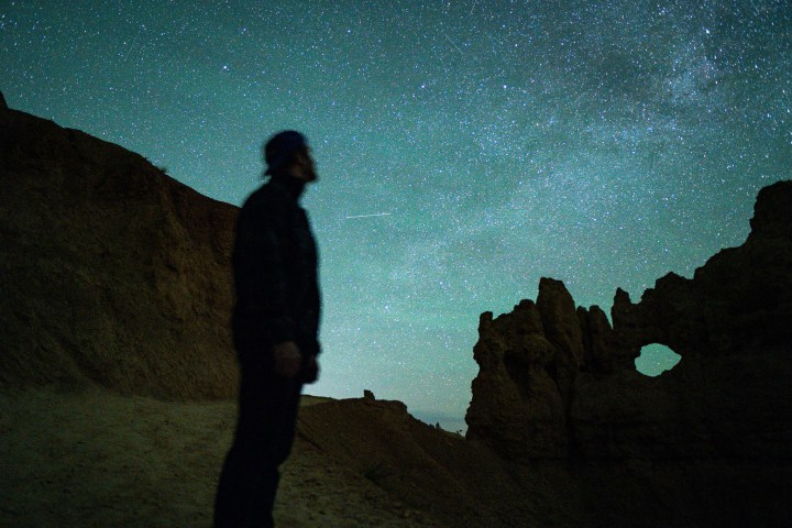 Silhouette of person stargazing near rock formations under starry sky.